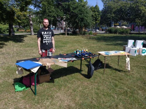 Ein Amnesty-Aktivist steht hinter einem Tisch im Stadthallenpark in Chemnitz. Auf dem Tisch liegen Farben und Farbrollen sowie Schablonen bereit, mit denen Stoffbeutel gestaltet werden können.